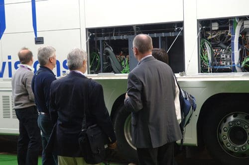 A group of five men examining the open compartment of a large vehicle, revealing complex machinery and wiring inside. They are engaged and focused, with a backdrop of a white vehicle featuring blue logos.