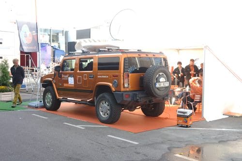 An orange Hummer SUV parked on an orange mat at a promotional event, with a tent and people gathered in the background. The atmosphere is lively, with a mix of attendees and displays around the area.