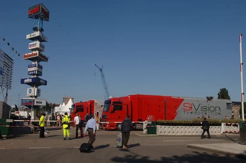 A busy scene at an event venue with several large, red television production trucks parked. People, including workers in bright safety gear, move around, and a tall sign and cranes are visible in the background under a clear blue sky.