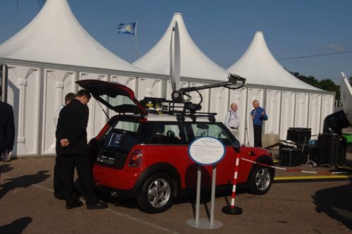 A red Mini Cooper with the rear hatch open, featuring a mounted communications antenna. In the background, people in suits stand beside white tents, engaged in conversation. The setting appears to be an outdoor event.