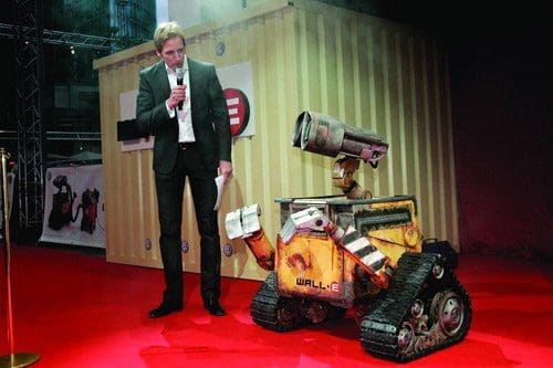 A man in a black suit holds a microphone while standing next to a small, animated robot named WALL-E, which has a rusty yellow body and tracks. They are on a red carpet, with a backdrop of a container and event decorations.