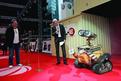 A robot character, Wall-E, stands on a red carpet at an event with two men, one holding a microphone, speaking to the audience. The background features banners and buildings, highlighting a festive atmosphere.