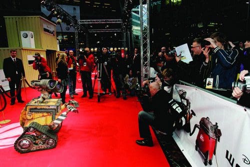 A small, animated robot with a rusty, yellow exterior posing on a red carpet, surrounded by photographers and fans. The crowd is capturing the moment with cameras while wearing event badges.