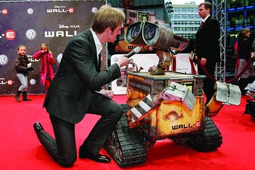 A man in a suit kneels beside a large, playful robot named Wall-E at a red carpet event, interacting with it. Children watch in the background, and a banner featuring Disney and Volkswagen advertisements is visible.