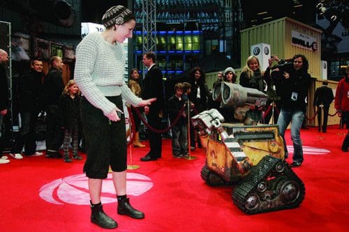 A woman wearing a sweater and shorts interacts playfully with a small robotic character on a red carpet event. Surrounding them are spectators and media capturing the moment, with a modern urban background visible.