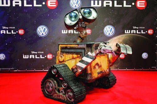 A robot character resembling Wall-E stands on a red carpet at an event. The robot has large, expressive eyes and a rusted, metallic body with tracks for mobility, set against a backdrop with the logos of Disney and Volkswagen.