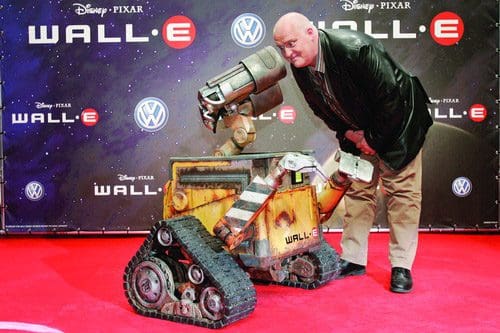 A person in a black jacket poses next to a life-sized WALL-E robot on a red carpet at a promotional event, with a backdrop featuring the Disney Pixar logo and a Volkswagen emblem.
