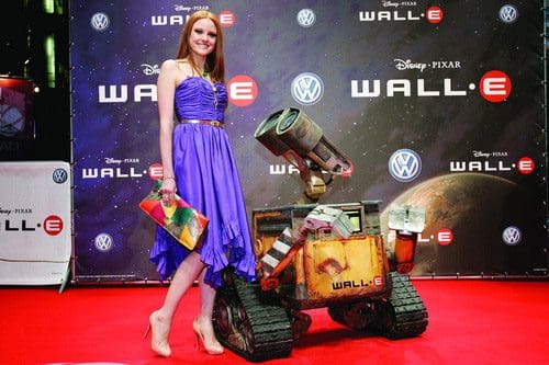 A woman in a purple dress stands beside a small robot character named Wall-E at a movie premiere event, with a red carpet and promotional banners in the background.