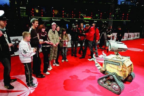 A small robot resembling Wall-E stands on a red carpet surrounded by a diverse group of people, including children and adults, some taking photos. The scene is lively with bright lights and a festive atmosphere.