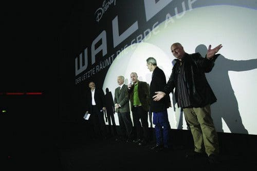 Four men standing on stage in front of a large screen displaying the movie title 'WALL-E.' The men, dressed in various outfits, appear to be engaged in a presentation or announcement related to the film.