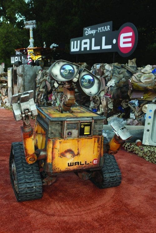 A small, charming robot named WALL-E, with large expressive eyes and tracks for movement, is placed on a red carpet. Behind him is a promotional display featuring the title 'WALL-E' by Disney-Pixar, and a backdrop made of various scraps and electronics.