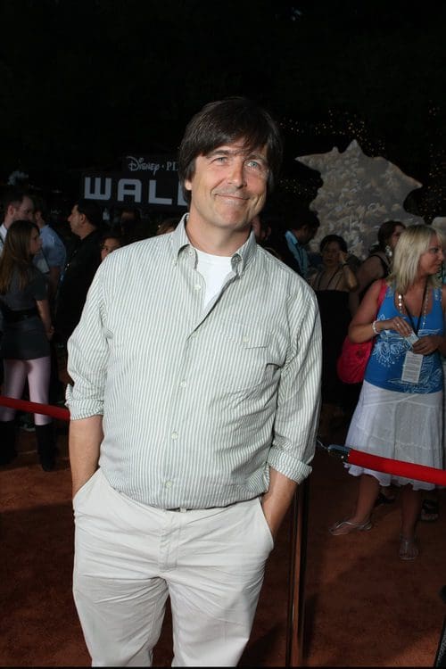 A man in a light green striped shirt and beige pants stands smiling on a red carpet at a lively event, surrounded by a crowd of people and colorful decorations in the background.
