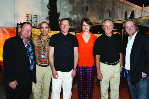 A group of six individuals standing together outdoors at an event. The men wear casual attire, while one woman in a bright orange top and patterned pants stands at the center, smiling. The backdrop features urban architecture and greenery.