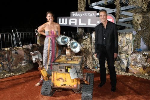A woman in a colorful striped dress and a man in a black suit stand beside the character Wall-E, a small yellow robot, at a movie premiere. The backdrop features the movie title 'WALL•E' against a textured, natural setting.