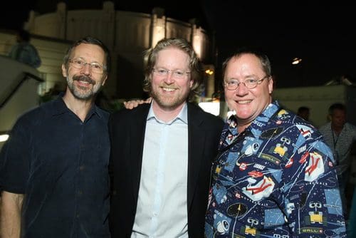 Three men standing together outdoors at night, smiling for the camera. The background features soft lighting and an illuminated building, creating a festive atmosphere. One man wears a dark shirt, the second a lighter one, and the third a colorful patterned shirt.