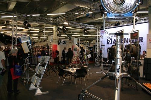 An exhibition space featuring various lighting equipment and structures, with people browsing booths in a spacious venue. A large spotlight is visible in the foreground, while banners and displays are set up in the background.