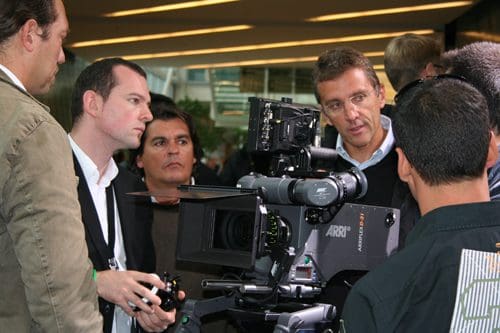 A group of professionals gathered around a film camera, discussing its features. The men display expressions of interest and concentration, with various backgrounds visible in a modern setting.