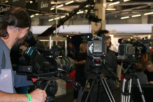 A man operating a video camera while another camera is set up on a tripod nearby. In the background, several people are engaged in conversation at a media event.