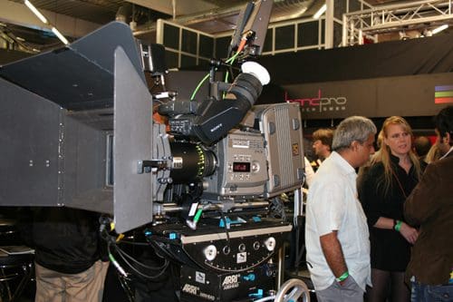 A professional filming camera on a wheeled tripod is prominently displayed at a trade event. Attendees, including a man in a white shirt and a woman in a dark blazer, engage in conversation in the background.