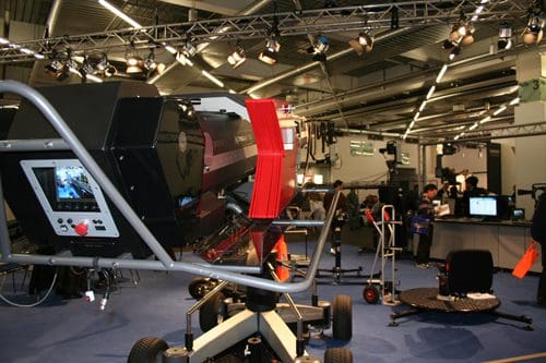 A large piece of advanced technology equipment, possibly for filming or surveillance, is showcased at a trade show. It features a display screen and a red component, with visitors and booths visible in the background.