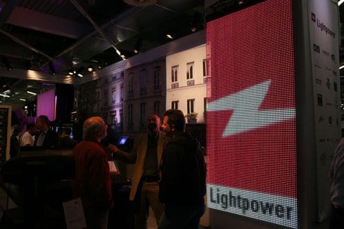 A group of attendees engaging in conversation at an exhibition booth with a large LED display showing the logo of Lightpower, in a well-lit environment.
