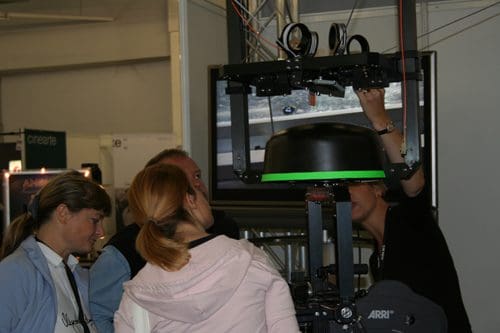 A group of three individuals observing a demonstration at a technology exhibition, focused on a sophisticated camera rig. A presenter explains the equipment, enhancing their interest in the setup.