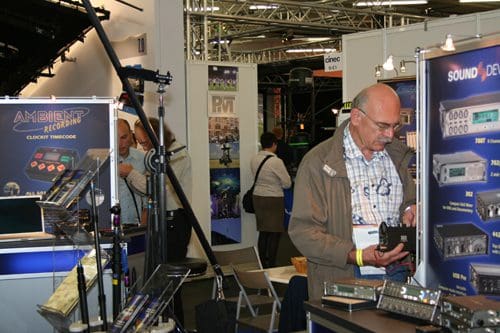 An older man wearing glasses inspects audio equipment at a trade show, surrounded by display booths featuring various sound gear and promotional materials, with other attendees visible in the background.