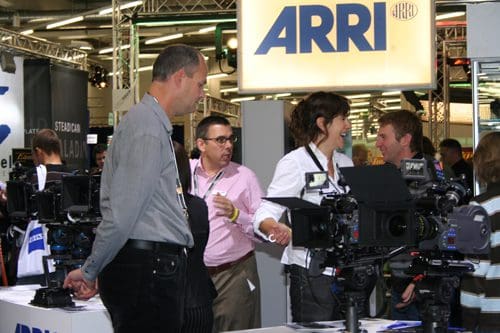 A group of professionals engaged in conversation at a film equipment exhibition. Various cameras are showcased on a table, with attendees discussing features and options in a bustling environment.
