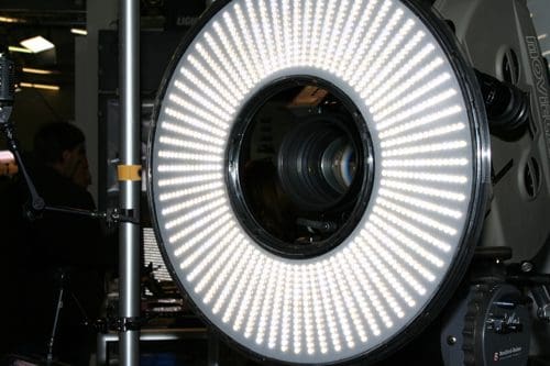 A close-up of a large circular LED light ring, showcasing bright, evenly distributed lights. The center includes a camera lens, and the surrounding area features equipment typical of a photography or film setup.