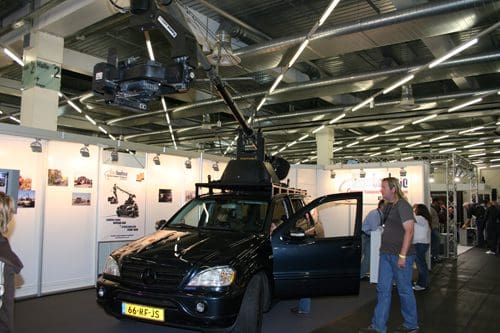 A black SUV with a mounted camera crane on its roof, parked in an exhibition hall. Attendees are walking nearby, with various display booths in the background showcasing media equipment.