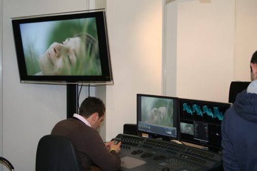 A film editing workstation featuring a seated editor focused on digital editing equipment. A large screen displays a close-up of a serene woman resting among grass, while additional monitors provide visual data.