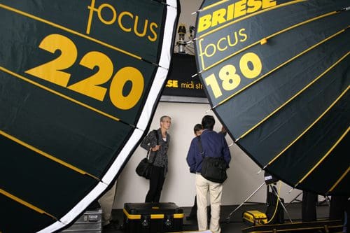 Two large softbox lights labeled "focus 220" and "focus 180" set up in a photography studio. A woman with short hair and a man in a dark jacket stand in front of a seamless backdrop, with photography equipment visible on the floor.