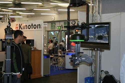 An exhibition booth showcasing film equipment, featuring a monitor displaying video footage and a camera on a stand. A person is observing the equipment in a bustling trade show environment.