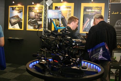 A professional video camera displayed on a circular table at a trade show, with bright blue lighting. Two attendees are talking behind the camera, while promotional posters are visible on the walls.