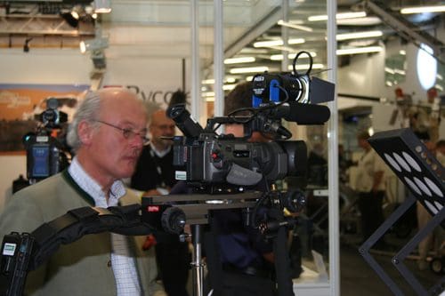 A man with glasses and a short white beard operates a professional video camera at an event. Behind him, other individuals and equipment are visible, suggesting a media or broadcast setting.