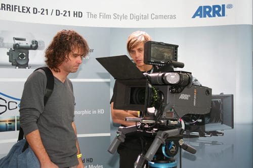 Two men discussing an ARRI digital camera at a film equipment booth. One man with curly hair inspects the camera, while the other, with straight blonde hair, points toward the equipment, engaged in conversation.