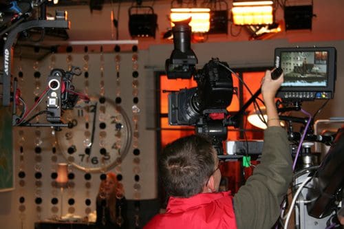 A person operating a camera in a studio, holding a monitor to check the shot. Soft lighting is set in the background, highlighting a large wall clock. Other cameras and equipment are visible, indicating a professional filming environment.