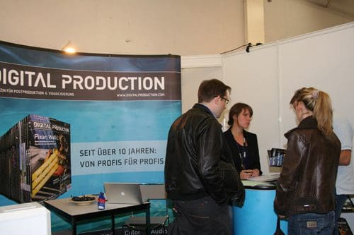 A booth displaying 'DIGITAL PRODUCTION' at an exhibition. A woman engages with two attendees, one male wearing glasses and a leather jacket, and a female in a light top. The booth features promotional materials and a laptop on the table.