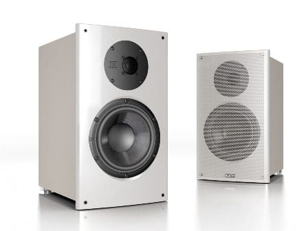 Two audio speakers displayed on a reflective white surface. The larger speaker on the left has a sleek design with a prominent woofer and a tweeter, while the smaller speaker on the right features a silver mesh front and a circular speaker design.