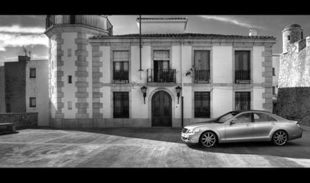 A sleek silver sedan parked in front of a grand, historic white villa with dark-framed windows and decorative lanterns, set against a cloudy sky. The image is presented in black and white, emphasizing the architectural details.