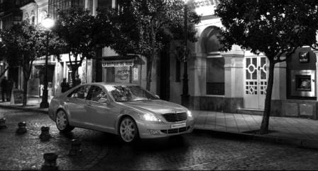 A sleek silver car parked on a cobblestone street at night, surrounded by trees. Soft streetlights illuminate elegant buildings in the background, creating a serene urban atmosphere.