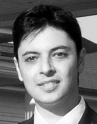 A man with short dark hair, wearing a black suit and white shirt, smiles for the camera. The image is in black and white, highlighting his facial features against a blurred background.
