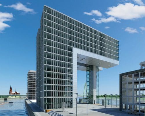 A modern glass building with a distinctive overhanging section, located by a waterfront. The structure features multiple glass panels reflecting the sky, with a nearby bridge visible in the background.
