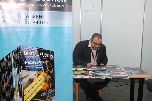 A man in business attire sitting at a table covered with magazines at a trade fair booth. Behind him is a large banner for 'DIGITAL PRODUCTION' showcasing various publications.