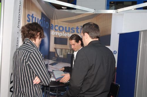 Three men are engaged in conversation at a trade show booth, discussing studio acoustic solutions. The backdrop features a modern studio design, promoting acoustic products and services. The setting conveys a professional environment.