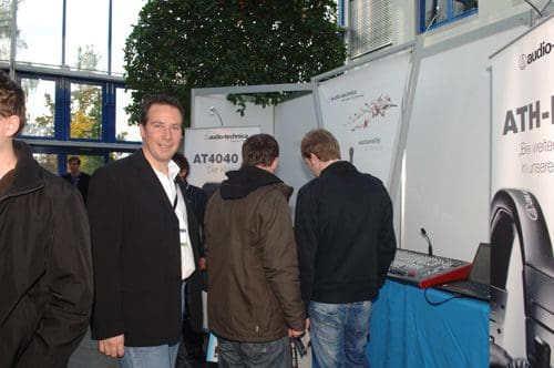 A man in a black jacket stands smiling in front of an audio equipment booth at an event. Two attendees in casual clothing are examining the displays, surrounded by plants and brightly lit interior space.