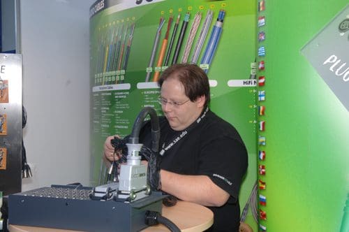 A man with glasses working at a table in an exhibition setting, focusing on a piece of equipment. Behind him, a green backdrop displays an array of colorful wires and technical information.