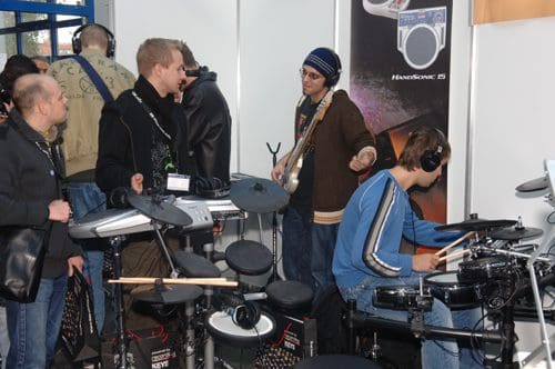 A group of people engaged in a music demonstration at a trade show, with one man playing an electronic drum set while others observe and discuss. The setting features various musical instruments and equipment.