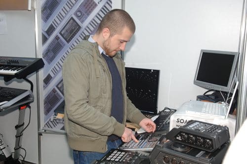 A man in a gray jacket interacting with electronic music equipment at a trade show. He stands in front of various audio devices and a computer monitor, focused on his task amidst a professional setup.