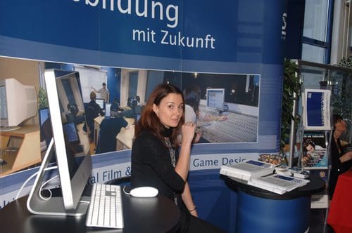 A woman with brown hair seated at a booth with a large computer monitor and keyboard. The backdrop displays images and text promoting educational programs in film and game design.